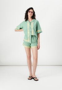 Groene linnen korte mouw shirt en bijpassende shorts met gele bloemenborduursels. Model draagt zwarte sandalen en paarse zonnebril.