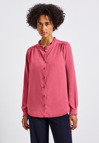 Blusa rosa a maniche lunghe con scollatura volante, chiusura a bottoni e cuciture delle spalle arricciate. Tessuto morbido, abbinata a pantaloni blu navy.