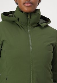 Chaqueta verde con cremallera y cuello alto, que presenta una textura suave y mate, con detalles en naranja en la herrajes.