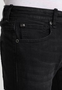 Los jeans de mezclilla negros presentan una vista en primer plano de la cintura, un pequeño bolsillo delantero y herrajes metálicos con el logotipo de la marca. Tela texturizada.