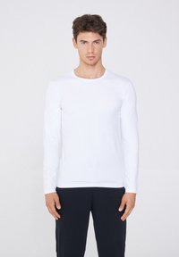Camisa blanca de manga larga hecha de tejido suave, con escote redondo. El modelo lleva pantalones oscuros y está de pie frente a un fondo neutro.