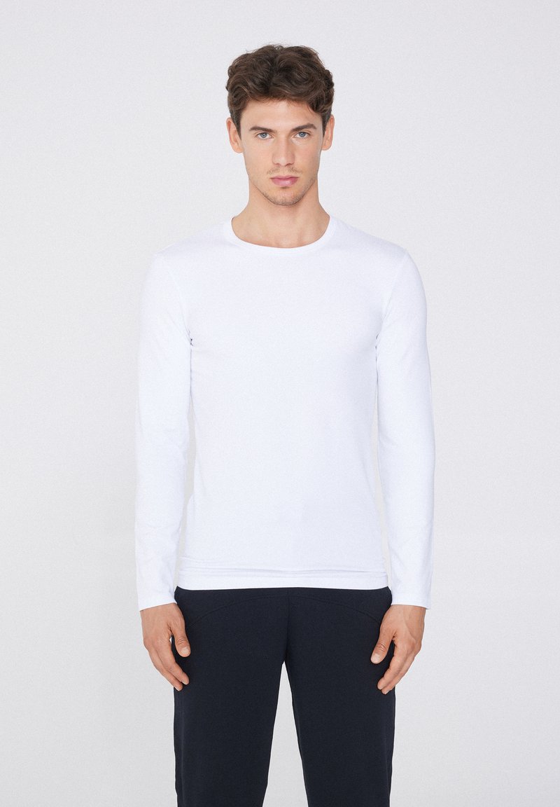 Camisa blanca de manga larga hecha de tejido suave, con escote redondo. El modelo lleva pantalones oscuros y está de pie frente a un fondo neutro.