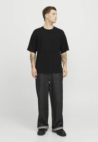 Camiseta negra de gran tamaño en tejido suave, combinada con pantalones de mezclilla oscuros y holgados con puños enrollados y costuras visibles. Footwear negro completa el look.