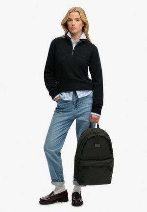 Vrouw staat met een zwarte trui met kwartrits, blauw gestreept overhemd, opgerolde spijkerbroek, witte sokken, donkere loafers, en houdt een zwarte rugzak aan het handvat vast.