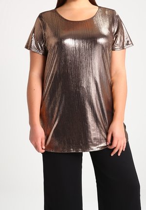 Kobieta ubrana w błyszczącą metaliczną, brązową bluzkę z krótkim rękawem i okrągłym dekoltem oraz czarne szerokie spodnie, stojąca na jednolitym tle.