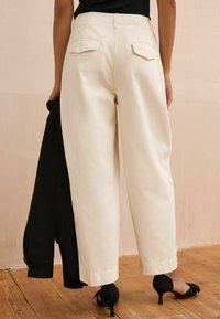 Pantalons larges couleur crème avec poches arrière, fabriqués en tissu doux. Le modèle tient un vêtement noir et porte des chaussures à talons noires.