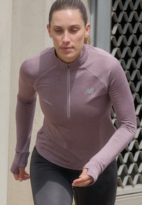Femme portant un haut de sport mauve zippé et un legging noir courant en extérieur devant un bâtiment avec une fenêtre à grille métallique.
