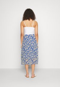 Gonna maxi floreale con base blu e fiori gialli, design a strati, tessuto leggero, abbinata con una canotta bianca aderente e sandali.