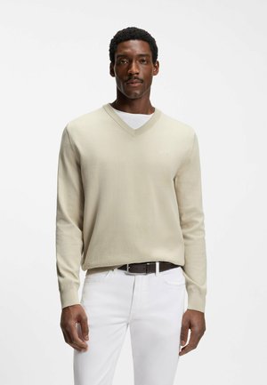 Mand iført beige V-hals sweater over hvid skjorte, hvide bukser og mørkt bælte, stående mod en ensartet hvid baggrund.