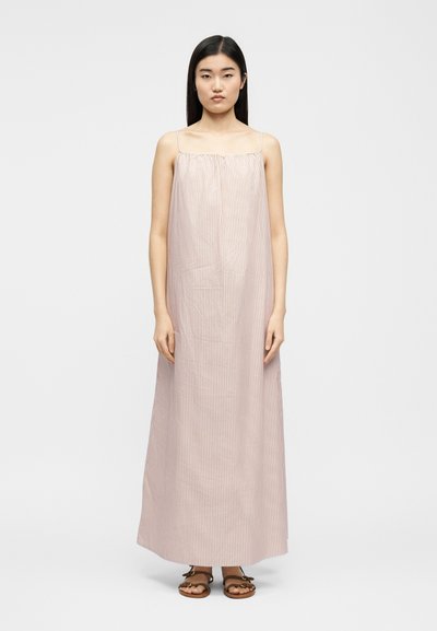 Mlada ženska stoji, oblečena v brezrokavno, do gležnjev dolgo svetlo roza obleko s tankimi navpičnimi črtami in rjavimi sandali.