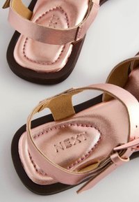 Roséguld metalliska sandaler med justerbara remmar och en brun sula, med sömdekorationer och präglad logotyp på innersulan.