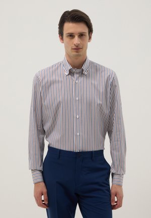 Hombre vistiendo una camisa de rayas de manga larga abotonada, metida dentro de pantalones azul marino, de pie contra un fondo claro y liso.