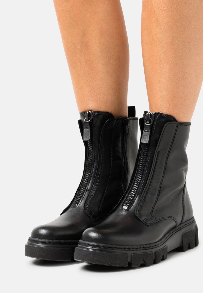 Bottes de cheville en cuir noir avec fermetures éclair à l'avant, portées sur des jambes nues, dotées de semelles épaisses et de languettes à l'arrière.