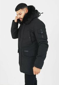 Svart parkasjacka med en huva foder av konstpäls, med flera fickor, dragkedja fram och en texturerad fabriksfinish.