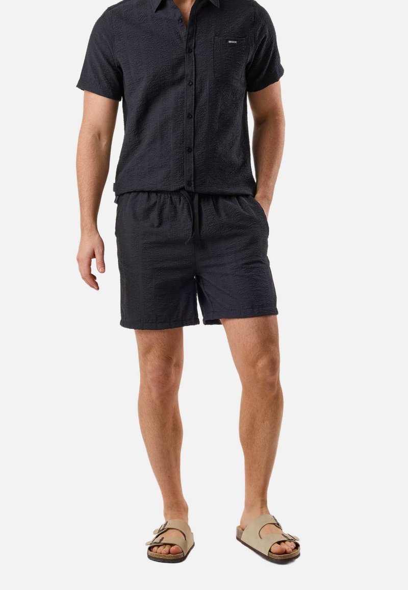Homme portant une chemise texturée noire à manches courtes avec boutons et un short assorti, ainsi que des sandales beiges à boucle, sur fond blanc.