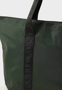 Bolsa tote de nailon verde con una correa negra gruesa que presenta letras embosadas, textura mate y un acento rectangular de tela negra en el punto de unión.