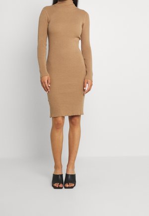 Mujer con un vestido ajustado de canalé de manga larga de color camel con cuello alto y zapatos mule negros con tacón y abertura en los dedos.