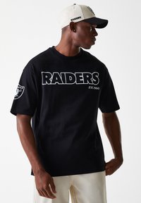 Juodas medvilninis marškinėliai su baltais užrašais "RAIDERS EST. 1960", su logotipo pleistru ant kairiojoRankovės. Dėvimas su smėlio spalvos ir juodu kepure.