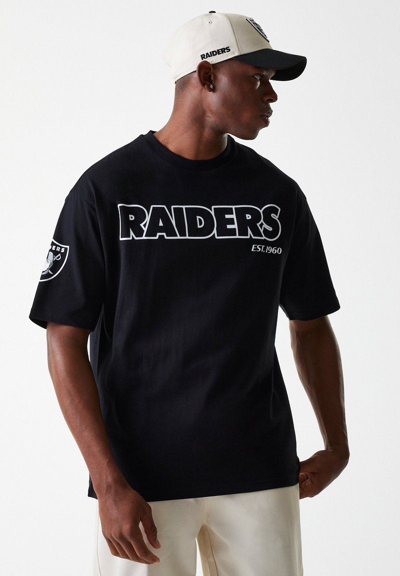 Juodas medvilninis marškinėliai su baltais užrašais "RAIDERS EST. 1960", su logotipo pleistru ant kairiojoRankovės. Dėvimas su smėlio spalvos ir juodu kepure.