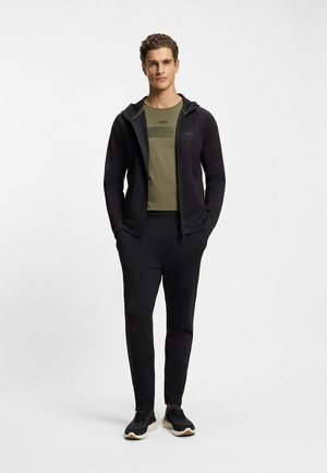 Mann trägt schwarzen Kapuzenpullover mit Reißverschluss, olivgrünes BOSS-T-Shirt, schwarze Jogginghose und schwarze Turnschuhe, steht mit den Händen in den Taschen.