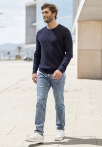 Marineblaue Rippstrickpulli, hellblaue Jeans und weiße Sneaker. Der Pullover hat einen runden Halsausschnitt und lange Ärmel; die Jeans haben einen geraden Schnitt.