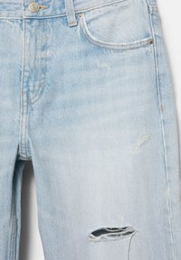 Ljusblå denimjeans med slitningar, synliga framfickor, metalldknappsstängning, bälteshällor och en trasig fläck nära knät.