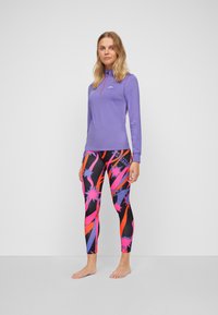 Helles lila Langarmoberteil mit hohem Kragen und pinkem Reißverschluss, kombiniert mit schwarzen Leggings, die ein leuchtendes abstraktes Design in Pink, Orange und Blau zeigen.
