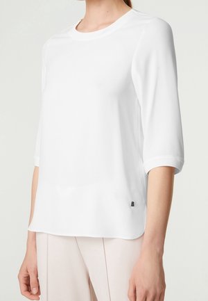 Vrouw draagt een effen witte top met driekwartmouwen en lichtbeige broek, staand tegen een witte achtergrond.