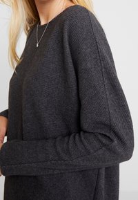 Ciemnoszary sweter o teksturowanej powierzchni z prążkowanej tkaniny, szerokim dekoltem i długimi rękawami, prezentowany na osobie z srebrnym naszyjnikiem.