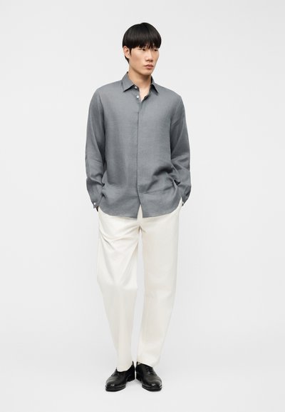 Άνδρας που φοράει χαλαρό γκρι πουκάμισο με κουμπιά, άσπρο παντελόνι με φαρδιά μπατζάκια και μαύρα παπούτσια, στέκεται με τα χέρια στις τσέπες μπροστά σε λευκό φόντο.