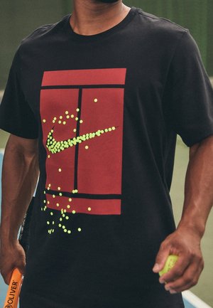 Mężczyzna w czarnej koszulce z czerwonym nadrukiem kortu tenisowego i żółtymi piłkami tenisowymi tworzącymi logo Nike, trzymający piłkę tenisową i rakietę.