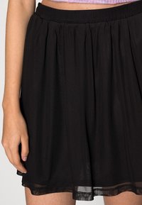 Jupe plissée noire avec une texture douce et transparente, dotée d'une taille élastique et d'un ourlet superposé légèrement volanté.