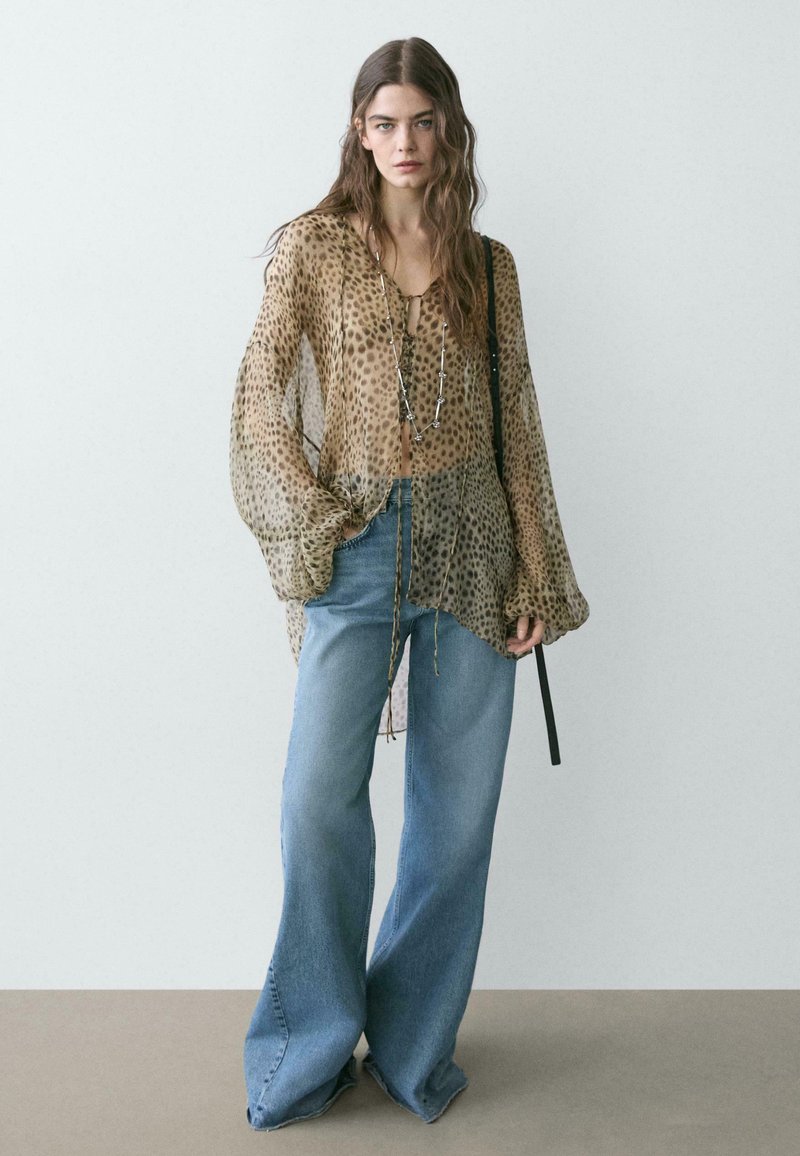 Kobieta w przezroczystej bluzce w panterkę, długim srebrnym naszyjniku, szerokich niebieskich dżinsach i z czarną torbą na ramię na jednolitym tle.