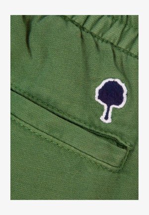 Green fabric with elastic stitching and a small embroidered dark blue tree with white outline near a pocket seam.