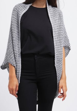 Femme portant un haut et un pantalon noirs avec un châle tricoté gris et léger drapé sur les épaules, sur un fond uni.