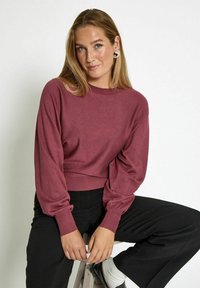 Maroon cropped sweater with long sleeves and ribbed cuffs, paired with black trousers. Smooth texture, minimal design, no patterns or embellishments.