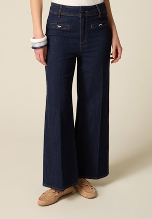 Kvinde iført mørkeblå jeans med høj talje og vide ben med frontlommer med klap og beige mokkasinsko, stående mod en ensfarvet baggrund.