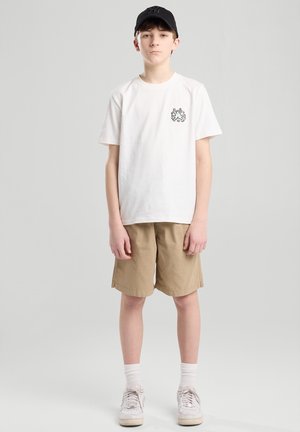 Witte katoen t-shirt met een kleine zwarte grafiek op de borst, gecombineerd met beige shorts en witte sneakers, staande tegen een grijze achtergrond.