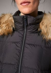Mujer vestida con una chaqueta de plumas negra con un cuello alto y un borde de piel sintética marrón alrededor de la capucha, con los labios visibles por encima del cuello.