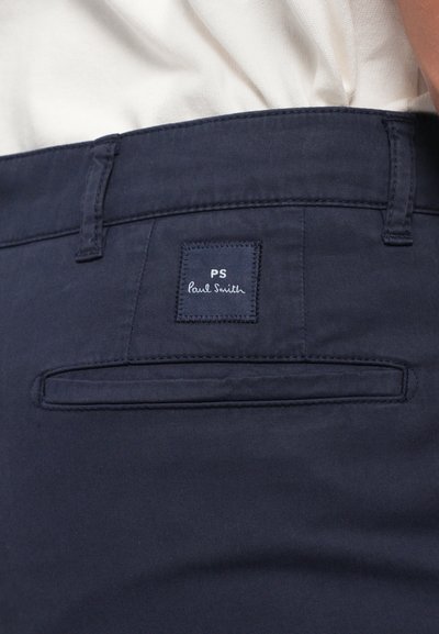 Pantaloni blu navy con tasca a filetto posteriore e etichetta quadrata nera con la scritta "PS Paul Smith" cucita sopra la tasca.
