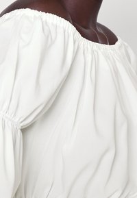 Blusa bianca off-the-shoulder con scollatura elastica arricciata e maniche voluminose, realizzata in un tessuto morbido e liscio con un leggero drappeggio.