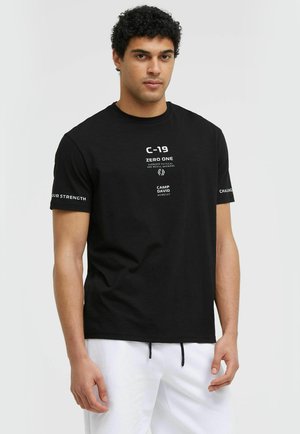 Man wearing a black graphic tee with text and white shorts, standing against a plain light gray background.