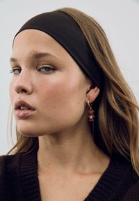 Brun stof hårbånd, lys brun sweater med ribbet tekstur, og hoops øreringe med en bjørn pendant og stjernedetalje. Nærbillede af ansigt.