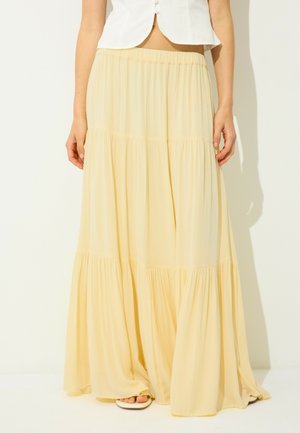 Femme portant un haut blanc sans manches et une jupe fluide jaune pâle, longue jusqu'aux chevilles, debout devant un fond clair et uni.