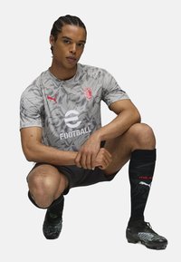 Grå kortærmet trøje med bladmønster, rød logo, sorte shorts og sorte sokker. Sorte og grønne fodboldstøvler med tekstureret detaljer.