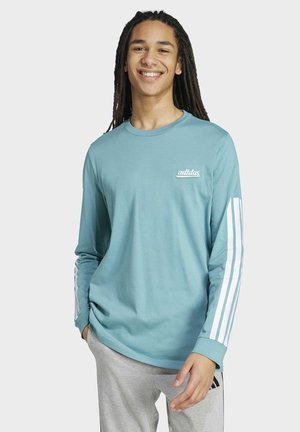 Persona sorridente con lunghe dreadlocks che indossa una maglia a maniche lunghe Adidas azzurro chiaro con strisce bianche sulle maniche e pantaloni grigi.
