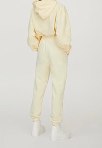 Jasnożółta, oversize'owa bluza z kapturem z kieszenią kangurką i sznurkiem, zestawiona z pasującymi dresami z elastycznymi mankietami. Białe sneakersy typu high-top.