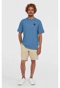 Blue cotton t-shirt with short sleeves and a round logo on the chest, paired with beige shorts and white sneakers.