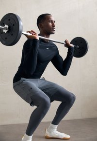 Atleta a realizar um agachamento com uma barra, vestindo uma camisola de manga longa preta, calções cinzas e leggings de compressão. Fundo em tons neutros.