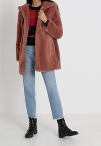 Kvinna som bär en brun faux pälsjacka, färggad tröja, ljusblå jeans och svarta nitar ankelskoro står mot en enkel bakgrund.